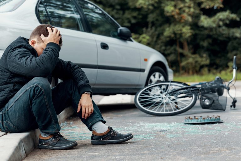 Potential concussion from biker holding his head and sitting on a pavement next to a car and bike crash