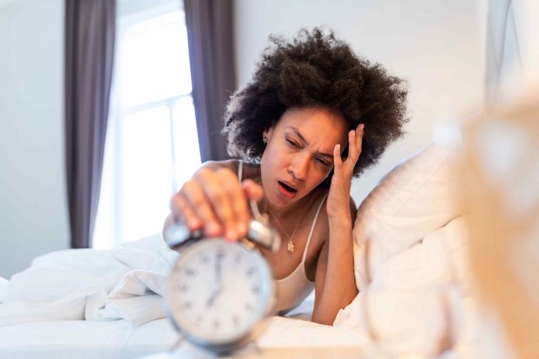 A fatigued ADHD adult waking up, reaching her alarm clock, illustrating the impact of daylight savings time in ADHD people.